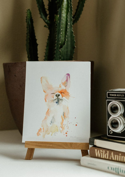 watercolor art of a proud and mischievous looking fox on a wooden stand, in front of a cactus plant next to a vintage camera.