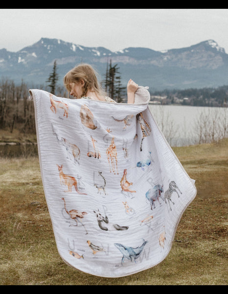 A girl outdoors in a grassy field with mountains and water in the background, holding up a white muslin blanket printed with alphabet animals and letters designed by Easy Sunday Club.