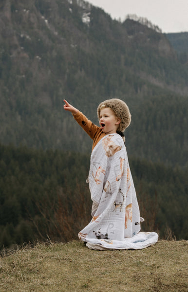 toddler wearing his raccoon hat and Alphabet Blanket as a cape in the mountains. He's pointing at a bird flying by and expressing his excitement.